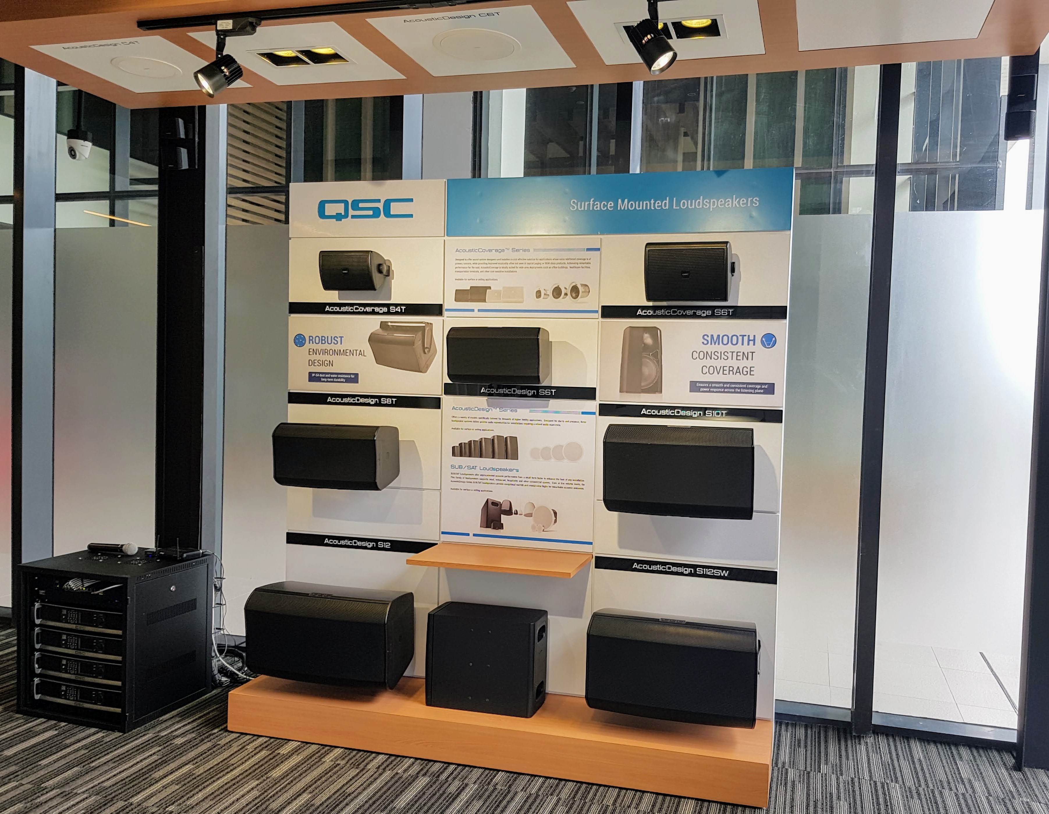 Display of various mounted Q-SYS speakers next to an AV rack equipped with QSC Amplifiers