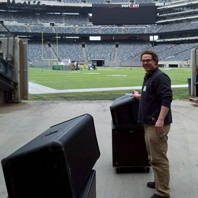 Don standing next to QSC loudspeakers next to the green of a football field